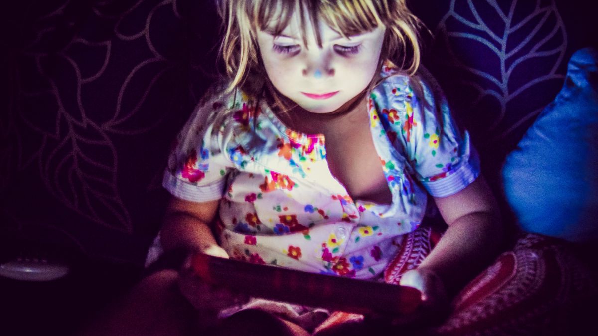4 year old girl using a tablet screen in a dark room with screen light illuminating her  on their face. Dark room. Night time. Impact of kids using devices in evenings at bedtime. blue light screen filter.