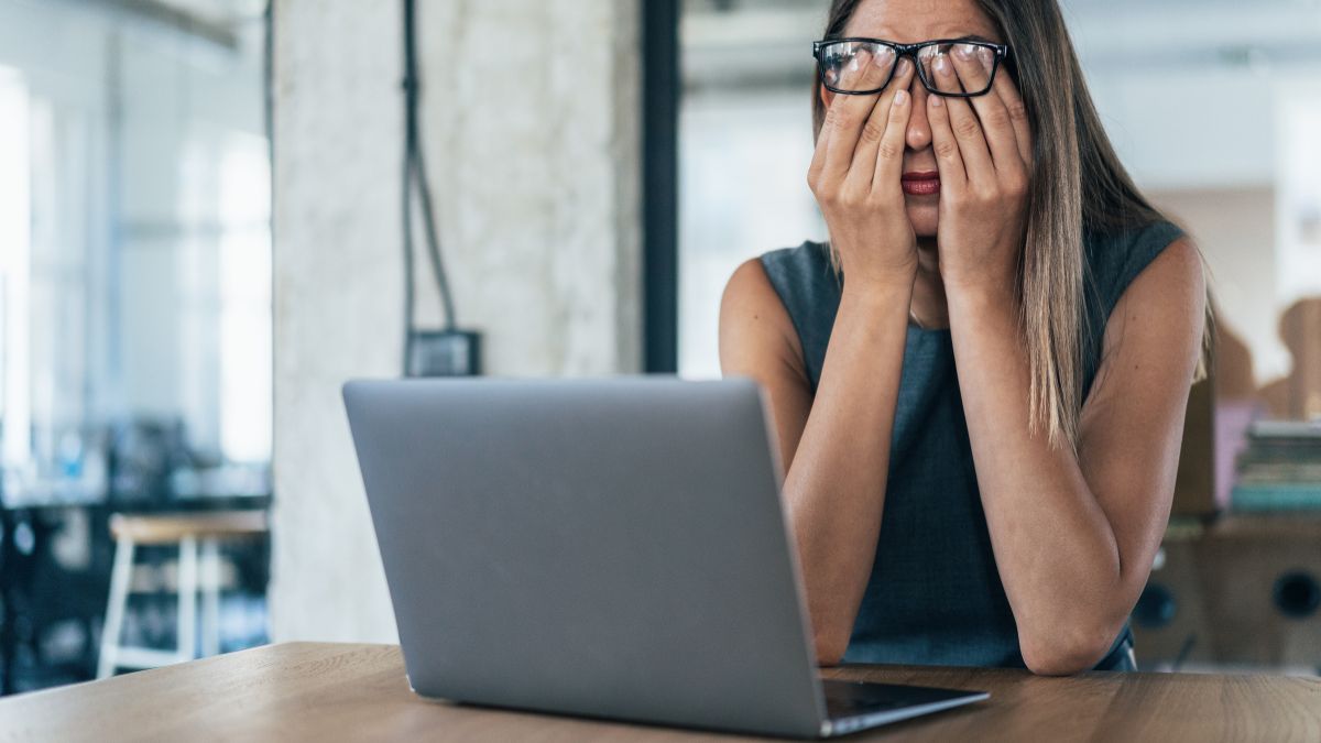 40 yr old Woman in an office rubbing tired eyes in front of a laptop screen after a long working day. She has glasses on her face which have moved up. daytime.
