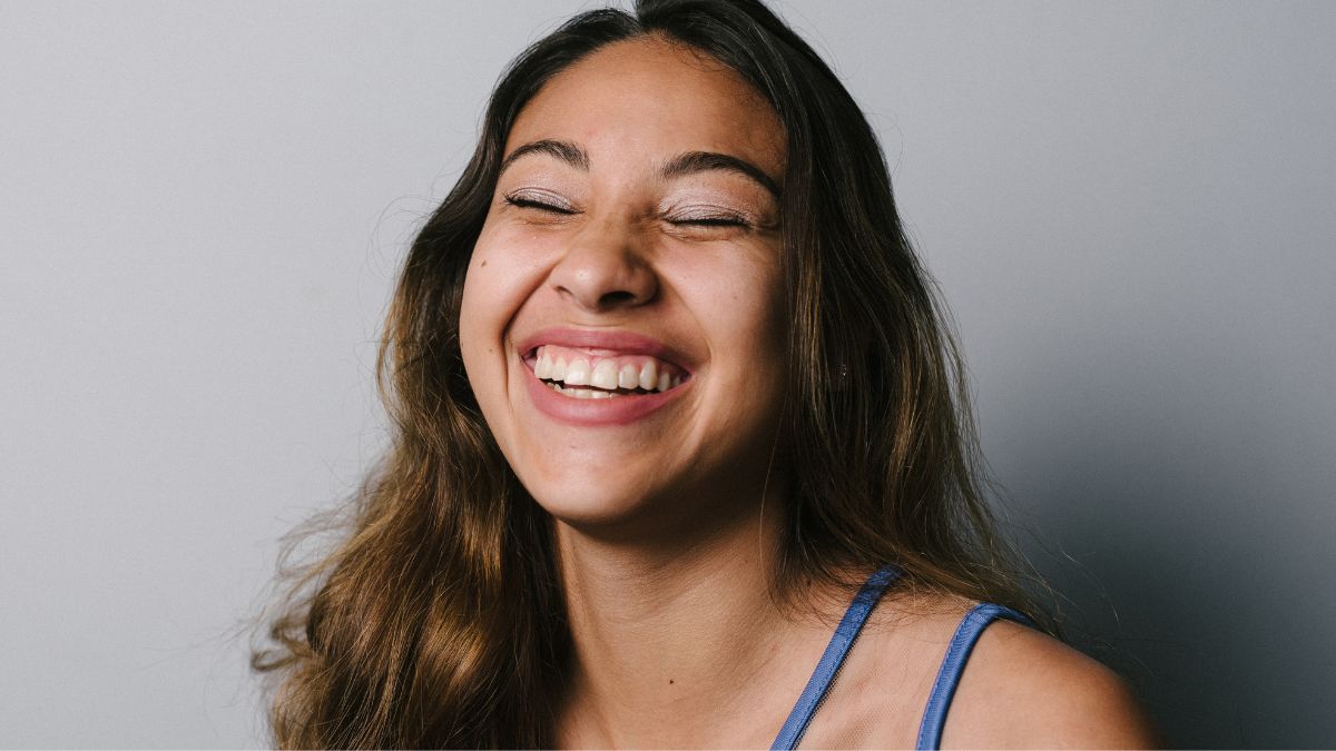 smiling 30yr old girl showing teeth. eyes closed, feeling joy and happiness. mild makeup on, plain light grey background. long brown hair.